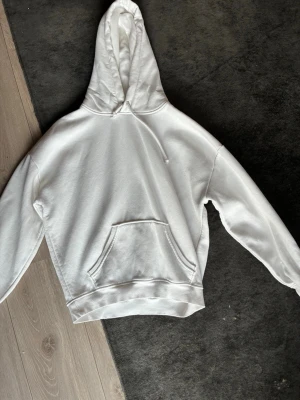 Vit hoodie med magficka och huva - Säljer en clean vit hoodie med stor magficka och dragsko i huvan. Hoodien har ribbade muddar vid ärmar och midja, samt en relaxed fit. Perfekt för dig som gillar enkel och stilren streetwear. Materialet känns mjukt och skönt mot huden.