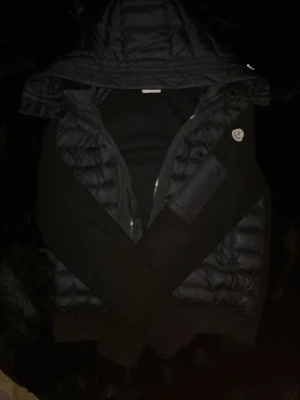 Svart dunväst med huva från Moncler - Snygg svart dunväst från Moncler med huva och dragkedja framtill. Västen har quiltad design och en Moncler-logga på bröstet. Perfekt för lager-på-lager och streetstyle. Materialet är glansigt dun och nylon, vilket ger en modern look.