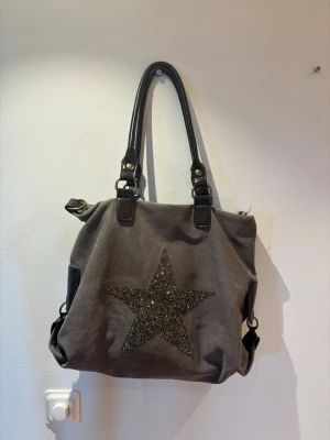 Grå handväska med glittrig stjärna - Snygg grå handväska i canvas med svarta detaljer och långa handtag i konstläder. Framsidan har en stor glittrig svart stjärna och baksidan en praktisk ficka med dragkedja. Väskan har metallspännen och sidofickor för extra förvaring.