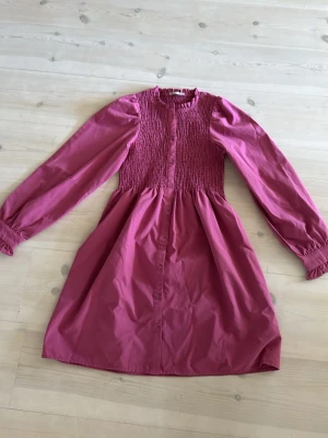 Rosa klänning med smock och knappar - Supersöt rosa klänning med smockad överdel och långa puffärmar. Klänningen har knappar framtill och är gjord i ett mjukt bomullstyg. Perfekt för dig som gillar en feminin och trendig stil.