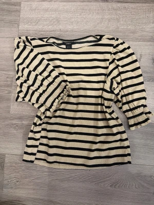 Randig topp med puffärm från Lindex - Säljer en beige och svart randig topp från Lindex med puffiga trekvartsärmar och resår vid ärmslut. Toppen har en lätt strukturerad yta och är gjord i ett mjukt material som känns skönt mot huden. Använt men fint skick. Lite nopprig.