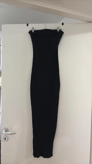 Svart ribbad axelbandslös klänning - Säljer en lång, svart axelbandslös klänning med ribbad struktur. Klänningen har en tight passform och är perfekt för dig som gillar minimalistisk och stilren design. Materialet känns mjukt och stretchigt, vilket gör den bekväm att bära.