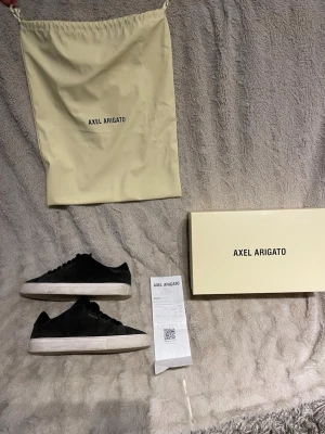 Svarta sneakers från Axel Arigato - Stilrena svarta sneakers från Axel Arigato i mocka med diskret guldfärgad logga på sidan. Skorna har vit sula och svarta skosnören. Kommer med originalkartong och dustbag. Perfekta för dig som gillar minimalistisk och clean design. 
