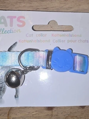  katt halsband med bjällra - Säljer ett reflekterande katt halsband från Cats Collection. Halsbandet är justerbart, har en blå spänne formad som ett kattansikte och en silverfärgad bjällra. Materialet är syntetiskt och bandet har en skimrande regnbågseffekt.