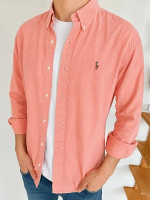 Korallfärgad skjorta från Ralph Lauren - Snygg korallfärgad skjorta från Ralph Lauren med klassisk button-down krage och broderad logga på bröstet. Skjortan har långa ärmar, vita knappar och är tillverkad i mjuk bomull. Perfekt att bära öppen över en t-shirt för en avslappnad look.