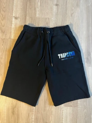 Svarta shorts med snörning - Svarta shorts från Trapstar med broderad logga i vitt och blått på vänster ben. Modellen har elastisk midja med snörning och två sidofickor. Perfekta för chill dagar och streetwear-stil.