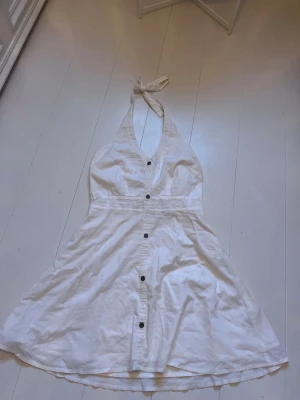 Vit halterneck klänning med knappar - Snygg vit halterneck klänning med knyt i nacken och knappar framtill. Klänningen har v-ringning och är ärmlös, perfekt för varma dagar. Tillverkad i mjuk bomull som känns skön mot huden och har en lätt utställd kjol.