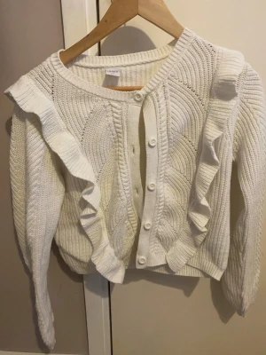 Vit kofta med volanger från Lindex - Säljer en vit stickad kofta från Lindex med dekorativa volanger längs framsidan och knappar hela vägen. Koftan har rund halsringning, lång ärm och ett snyggt mönsterstickat tyg. Perfekt för dig som gillar en söt och trendig stil.