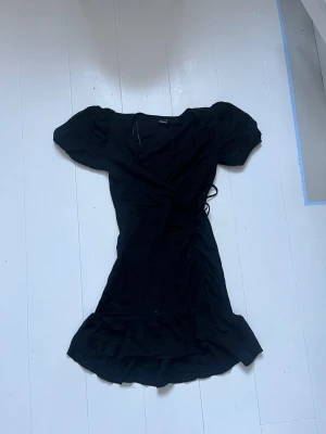 Svart omlottklänning med volang - Snygg svart omlottklänning med korta puffärmar och volangkant nedtill. Klänningen har knyt i midjan och är tillverkad i ett mjukt tyg som ger en skön passform. Perfekt för dig som gillar en enkel men trendig look.