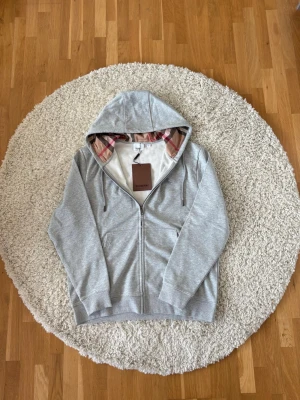Grå Burberry zip-hoodie   - Snygg grå hoodie med dragkedja från Burberry. Klassisk huva med det ikoniska rutmönstret på insidan, två fickor framtill och ribbade muddar. Perfekt för en chill och stilren look. Hör av er om ni vill ha fler bilder på detaljer osv. Pris kan diskuteras!