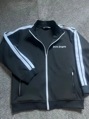 Palm Angels svart track jacket - Palm Angels svart track jacket med vita ränder längs ärmarna och vit dragkedja. Tröjan har hög krage, två fickor med dragkedja och logga på bröstet. Sportig och trendig design som passar perfekt till streetwear-stilen.