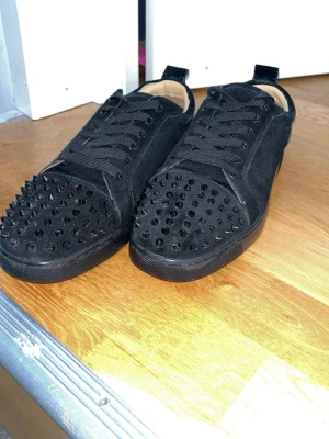 Christian Louboutin skor  - Säljer ett par svarta sneakers med coola svarta nitar på tån. Skorna har snörning, platt sula och är gjorda i mockaliknande material. Insidan är beige och de har en modern, edgy look som sticker ut.