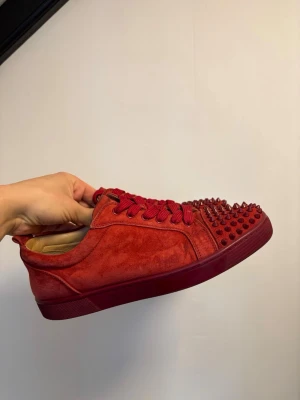 Röda Christian Louboutin jr sneakers  - Snygga röda sneakers från Christian Louboutin i mocka med matchande röda skosnören och coola nitar på tån. Skorna har en låg silhuett och röd gummisula som ger en riktigt unik vibe. Perfekta för dig som vill sticka ut med din stil.