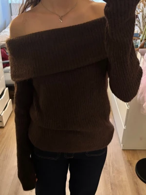 Brun off shoulder stickad tröja - Mysig brun stickad tröja med off shoulder-design och bred krage. Tröjan har långa ärmar och är ribbstickad för en snygg och avslappnad look. Perfekt för att hålla värmen och samtidigt se trendig ut.