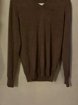 Beige V-ringad tröja J.Lindeberg - Stickad brun tröja från J.Lindeberg med V-ringning och diskret logga på bröstet. Tillverkad i mjukt material, perfekt för lager på lager. Klassisk passform med ribbade muddar vid ärmslut och nederkant.