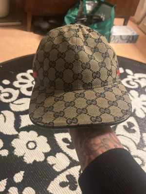 Gucci beige keps med GG-mönster - Säljer en beige keps från Gucci med det klassiska GG-monogrammet över hela kepsen. Kepsen har böjd skärm, justerbart spänne bak och detaljer i rött och grönt på sidorna. Materialet är canvas med läderdetaljer. Perfekt för dig som vill ha en lyxig och trendig accessoar.