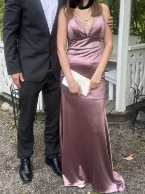 Lång lila satinklänning från Amarà - Elegant långklänning i skimrande lila satin från Amarà. Klänningen har djup v-ringning, smala axelband och figurnära passform som ger en snygg siluett. Perfekt för skolbal eller andra speciella tillfällen. (Pris kan diskuteras, köptes för 5600kr) använd en gång