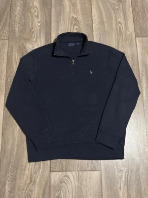 Mörkblå stickad tröja Polo Ralph Lauren - Mörkblå stickad tröja från Polo Ralph Lauren med halv dragkedja och klassisk broderad logga på bröstet. Tröjan har lång ärm, hög krage och ribbade muddar. Perfekt för lager på lager och en stilren look.