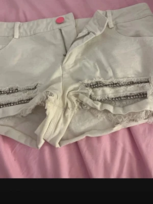 Vita shorts med slitningar och pärlor - Snygga vita shorts med slitna detaljer och pärlor längs framsidan. De har en rosa knapp, dragkedja och råa kanter nertill. Perfekta för sommarens alla äventyr och ger en cool vibe till din outfit.