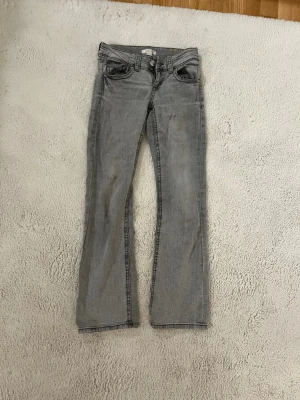 Grå bootcut jeans från Gina Tricot! - Säljer ett par grå bootcut jeans från Gina Trickot med klassisk femficksdesign och silverfärgade nitar. Jeansen har låg midja och är tillverkade i mjuk denim med lätt tvättad look. Perfekta för dig som gillar en avslappnad men ändå trendig stil.