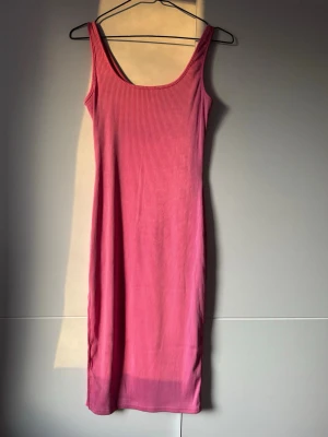 Rosa ribbad långklänning - En enkel och snygg rosa långklänning med ribbad struktur. Klänningen är ärmlös med breda axelband och har en rak, kroppsnära passform. Perfekt för varma dagar eller att styla med en jacka. Materialet känns mjukt och stretchigt.