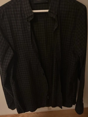 Mörkgrå rutig skjorta - Snygg mörkgrå skjorta med diskret rutigt mönster. Klassisk krage och långa ärmar, perfekt för en stilren look. Skjortan har knappar framtill och är tillverkad i ett mjukt bomullstyg som känns bekvämt hela dagen. Priset går att diskutera!!