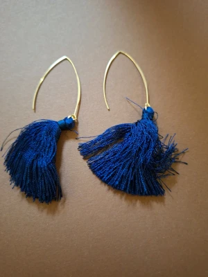Blå tofsörhängen med guldfäste - Snygga örhängen med mörkblå tofsar i textil och tunna, böjda guldfärgade metallbågar. Perfekta för dig som vill ha en färgklick och en lekfull touch till din stil. Lätta att bära och ger ett trendigt intryck.