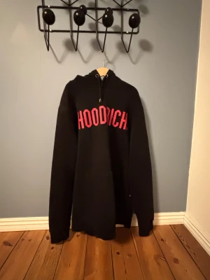 Svart hoodie från Hoodrich med tryck - Svart hoodie från Hoodrich med stor röd logga över bröstet. Klassisk huva med dragsko och känguruficka framtill. Tillverkad i mjukt material som passar perfekt för en chill och avslappnad stil.