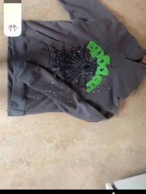 Grå spider hoodie  - Grå hoodie med huva och tryck av ett stort svart spindelnät på bröstet samt grön text som säger 'spooky'. Ärmen har vita och gröna färgstänk för en extra cool vibe märket är även väll kändt i usa pris kan diskuteras vid snabb affär 