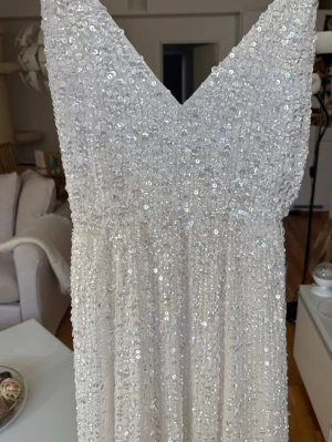 Vit långklänning med paljetter - Magisk vit långklänning täckt av glittriga paljetter. Klänningen har v-ringning och smala axelband, vilket ger en elegant och festlig look. Perfekt för dig som vill sticka ut och glänsa på nästa stora event.