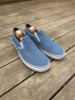 Blå slip-on sneakers från Vans - Säljer ett par klassiska blå slip-on sneakers från Vans. Skorna har en ovandel i canvas, vit sula med svart rand och den ikoniska Vans-loggan bak på hälen. Perfekt för dig som gillar enkel och stilren design.