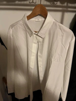 Vit klassisk skjorta i bomull - Stilren vit skjorta i storlek L med klassisk krage och knappar framtill. Skjortan har lång ärm och är tillverkad i mjuk bomull, perfekt för en clean och enkel look. Passar dig som gillar minimalistisk stil.