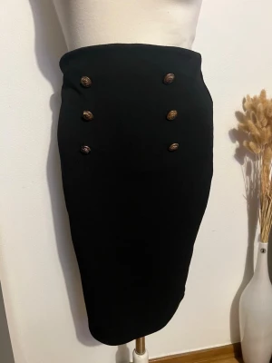 Svart pennkjol med guldknappar - Snygg svart pennkjol med sex dekorativa guldknappar framtill. Kjolen har en klassisk, figurnära passform och är perfekt för en stilren look. Materialet känns som polyester och kjolen har en hög midja som framhäver siluetten.