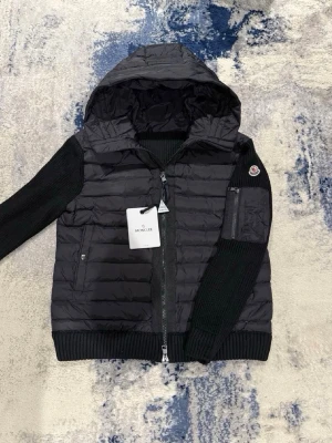 Svart cardiganjacka från Moncler - Snygg svart cardiganjacka från Moncler med vadderad framsida och huva i glansigt material. Ärmar och rygg är stickade i svart, med ribbade muddar. Dragkedja framtill och fickor med dragkedja. Moncler-logga på ena ärmen.
