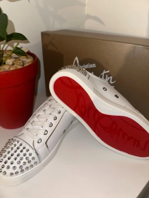 Vita Christian Louboutin sneakers med nitar - Säljer ett par vita sneakers från Christian Louboutin med ikoniska röda sulor och silverfärgade nitar på tå och häl. Skorna har vita skosnören och är tillverkade i slätt läder. Perfekta för dig som vill sticka ut med en lyxig och edgy stil.