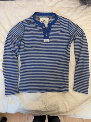 Blåvit randig långärmad t-shirt Hollister - Snygg långärmad t-shirt från Hollister i blått och vitt med smala ränder. Tröjan har tre knappar vid halsen och en liten etikett med Hollister-logga framtill. Tillverkad i mjuk bomull, perfekt för en avslappnad stil.