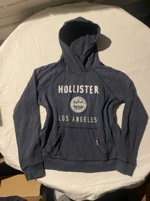 Mörkblå Hollister hoodie med tryck | 40 - Mörkblå hoodie från Hollister med stor huva och magficka. Framtill står det 'HOLLISTER LOS ANGELES' i vitt tryck och ett runt emblem med palmer och texten 'California Since 1922'. Mjuk bomullskänsla och ribbade muddar vid ärmar och nederkant.