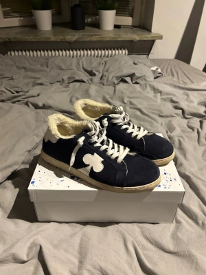 The Gallerist  - Snygga mörkblå sneakers med vit sula och vit klöverdetalj på sidan. Skorna har vita skosnören och är fodrade med mjuk fuskpäls för extra komfort. Perfekta för dig som vill sticka ut med en unik detalj och hålla fötterna varma. Har knappt använt dom har använt dom nångra få gånger.