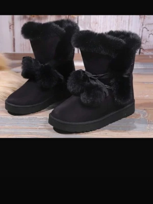 Svarta vinterboots med fuskpäls - Säljer ett par svarta boots med fluffig fuskpäls runt skaftet och stora pälstofsar framtill. Skorna har en mjuk ovandel och dekorativa rosetter. Perfekta för kalla dagar när du vill ha både stil och värme.