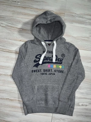 Grå Superdry hoodie med tryck - Säljer en grå hoodie från Superdry med stor logga och japansk text på bröstet🤩 Hoodien är i fint skick och passar Xs-S🙌🏻 Hör av er vid funderingar samt snabbaffär😇