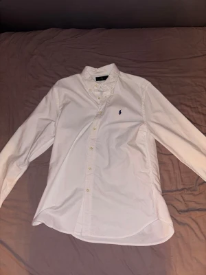 Vit skjorta från Ralph Lauren - Klassisk vit skjorta från Ralph Lauren med button-down krage och broderad logga på bröstet. Skjortan har långa ärmar, knappar framtill och är tillverkad i bomull för en skön och stilren look.