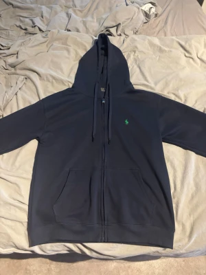 Mörkblå hoodie från Polo Ralph Lauren - Snygg mörkblå hoodie från Polo Ralph Lauren med dragkedja, huva med dragsko och klassisk grön logga på bröstet. Mjuk bomullskänsla och känguruficka framtill. Har använt den typ fem gånger.
