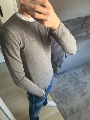 Grå stickad tröja från J.Lindeberg - Snygg grå stickad tröja från J.Lindeberg med diskret logga på bröstet. Tröjan har rund halsringning, ribbade muddar och är långärmad. Perfekt att matcha med jeans för en clean look.