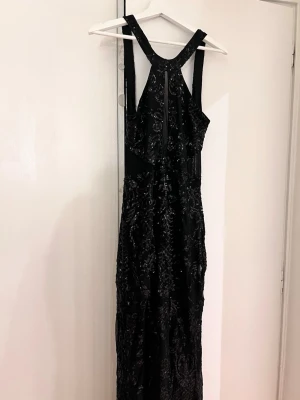 Svart långklänning med paljetter - Elegant svart långklänning med glittriga paljettdetaljer och meshpartier. Klänningen har breda axelband och en hög halsringning med en liten öppning framtill. Perfekt för dig som vill sticka ut på festen.