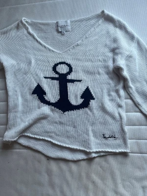 Vit stickad tröja med marinblå ankar - Vit stickad tröja från något oläsligt märke med ett stort marinblått ankare på framsidan. Tröjan har v-ringning och långa ärmar. Perfekt för en avslappnad och nautisk look. Materialet känns mjukt och lätt, vilket gör den skön att bära. Storleken är m men hade passat på S