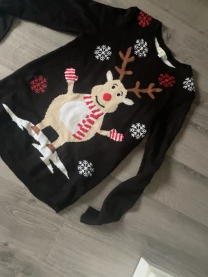 Svart jultröja med renmotiv  - Stickad svart jultröja med stort motiv av en ren med röda horn och röd-vit halsduk. Tröjan har snöflingor i vitt och rött samt långa ärmar och rund hals. Perfekt för julmys och vinterhäng.