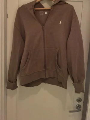 Brun hoodie från Polo Ralph Lauren - Säljer en brun hoodie från Polo Ralph Lauren med dragkedja, huva och känguruficka. Klassisk vit logga broderad på bröstet. Tillverkad i mjukt bomullsmaterial och har ribbade muddar vid ärmslut och nederkant. Perfekt för en chill och avslappnad stil.