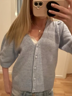 Ljusblå kofta med puffärm - Säljer en ljusblå kofta med v-ringning och knappar framtill. Koftan har korta puffärmar och ribbade kanter vid ärmslut och nederkant. Perfekt att slänga över en topp för en chill look. Materialet ser ut att vara mjukt och stickat.