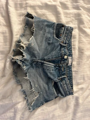 Blå jeansshorts med fransig kant Zara - Säljer ett par blå jeansshorts från Zara med hög midja och rå, fransig kant. Klassisk femficksmodell med dragkedja och knapp framtill. Perfekta för sommaren och ger en avslappnad vibe.