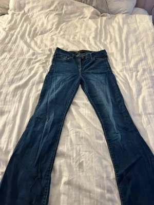 Blå bootcut jeans med midwaist - Säljer ett par klassiska blå bootcut jeans med låg/mid midja och fem fickor. Jeansen har en snygg tvätt och är tillverkade i stretchigt denimtyg för skön passform. Perfekta för dig som gillar retrovibbar och vill ha ett par jeans som sticker ut lite extra.